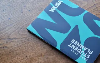 A 2020-2021 student planner with a blue and black cover labeled "WUSA Handbook" lies on a wooden table.