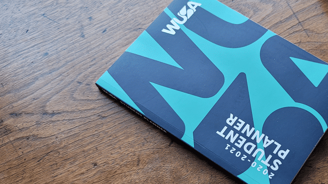 A 2020-2021 student planner with a blue-green and black cover labeled “Free WUSA Handbook” lies on a wooden table. The cover design features bold, abstract shapes. The planner is positioned slightly diagonally on the table. A 2020-2021 student planner with a blue and black cover labeled "WUSA Handbook" lies on a wooden table.