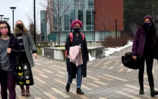 Four people, all wearing masks, walk on an outdoor path near a modern building at the University of Waterloo. They are dressed in winter clothing. One person is carrying a bag with a Batman logo, seemingly cautious amidst the ongoing WUSA outbreak affecting local residences.