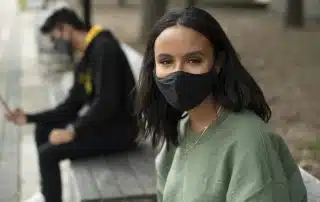 Two individuals sit on benches outdoors, both wearing black face masks. The person in the foreground is looking at the camera, while the other person in the background is pre-registering for a COVID-19 vaccine on their device.