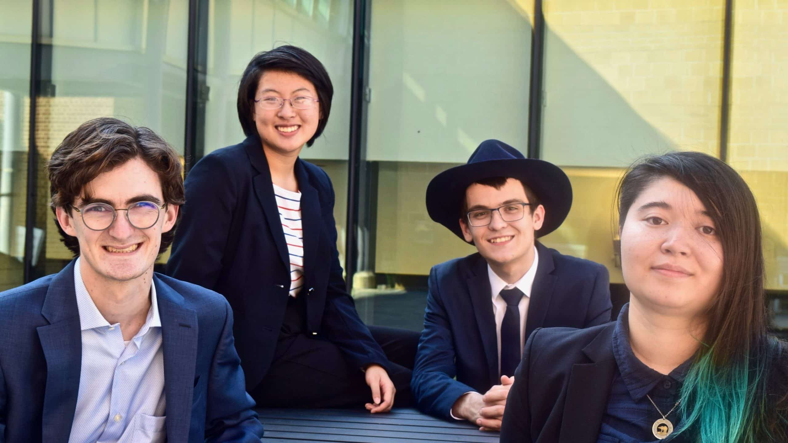 Four people wearing formal attire are sitting and posing for a group photo in an outdoor setting with glass-paneled walls in the background.