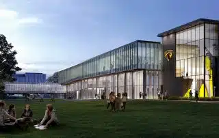 A modern glass and brick building with people sitting and walking on a green lawn in front, featuring a university logo on the building.