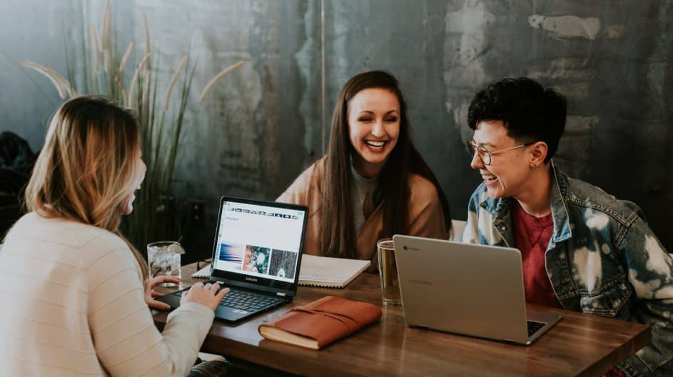 Three people are sitting at a wooden table with laptops and notebooks, engaged in conversation and smiling.