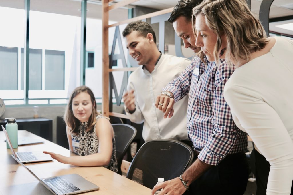 Four people engage in discussion around a table with laptops. Two men and two women appear focused and collaborative in a bright office setting.