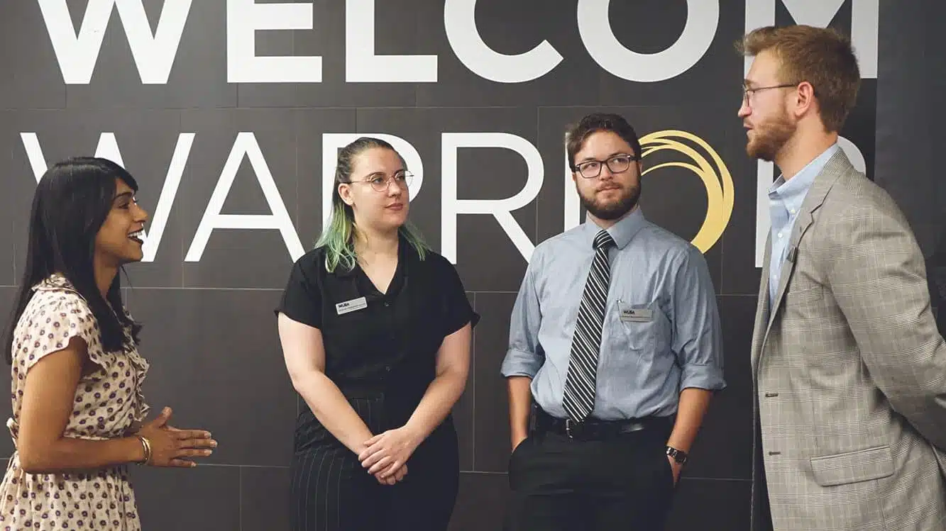 Four people in business attire have a conversation in front of a wall with the words "Welcome Warrior" on it.