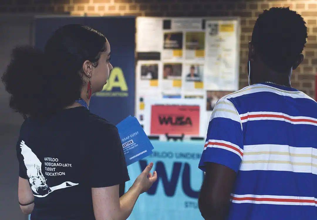Two individuals stand in front of a bulletin board covered with flyers and posters. Both are engaged in conversation about general meeting rules, with one holding brochures. The setting appears to be an indoor event or information booth.