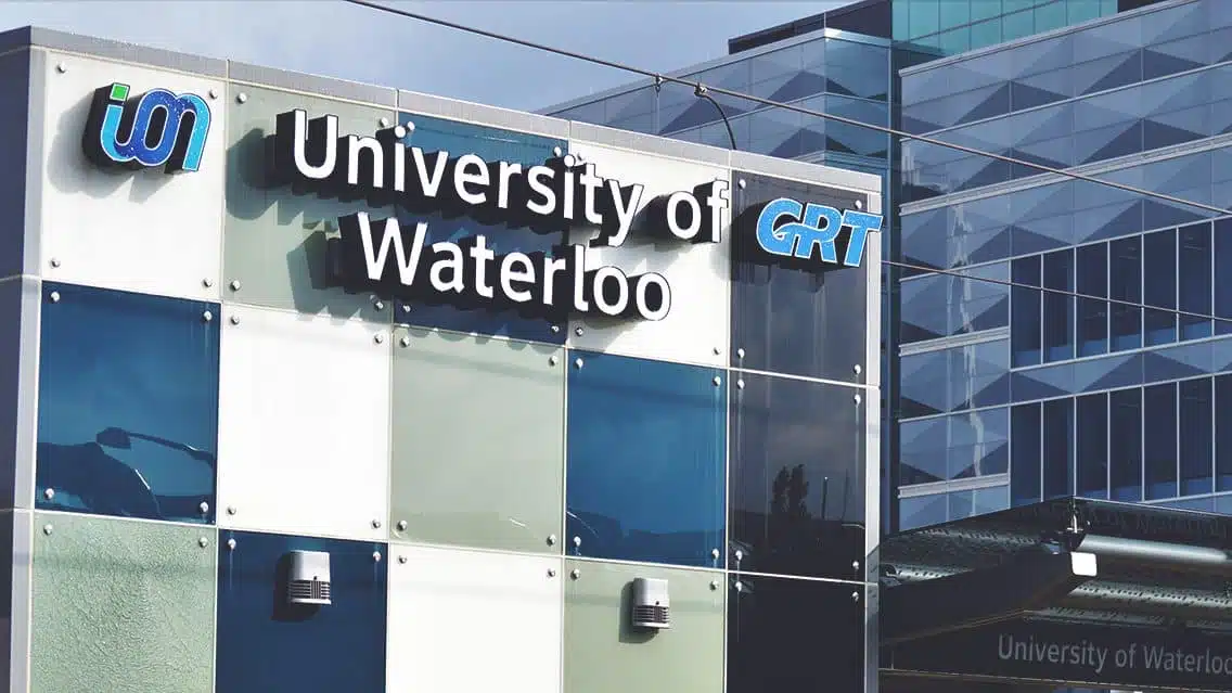 A sign for the University of Waterloo next to logos for local transit services U-Pass and GRT. The background includes parts of modern glass buildings.