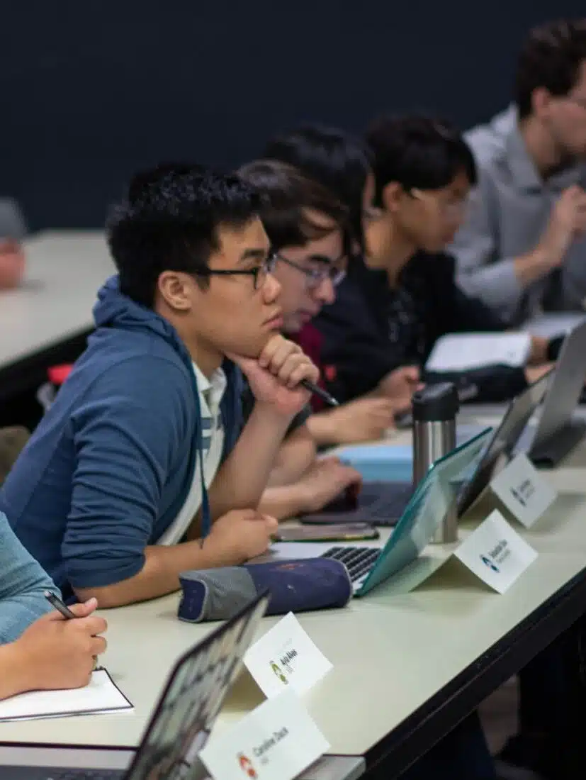 Students sitting at desks, adhering to general meeting rules, focus on their laptops and notepads during a classroom session.