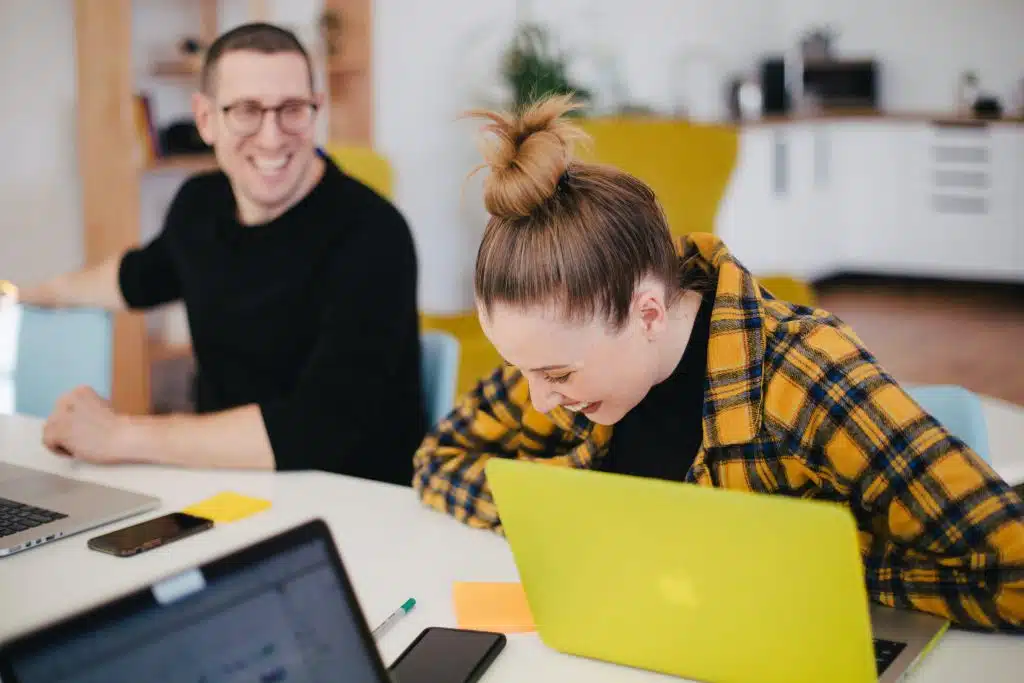 Two people are seated at a table with laptops, engaging in a conversation and smiling. One person wears glasses and a black shirt; the other has a bun and a yellow plaid shirt.