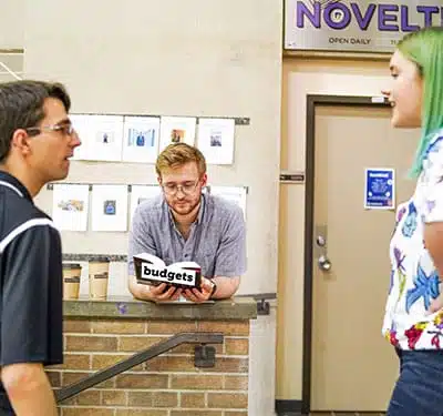 Three people are conversing indoors. One person holding a book labeled "budgets" is leaning over a brick barrier, while the other two, possibly discussing meeting guidelines, stand facing each other. A door and posters are in the background.