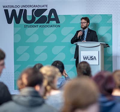 A person in a suit speaks at a podium labeled "WUSA" in front of an audience. The backdrop reads "WUSA Student Association General Meeting.