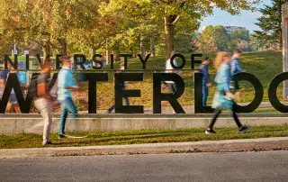 Students walk past a large "University of Waterloo" sign on a campus with trees and a grassy area in the background.