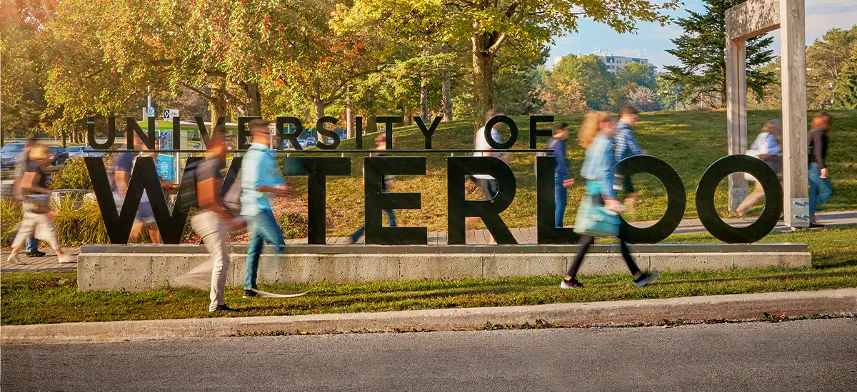 Students walk past a large "University of Waterloo" sign on a campus with trees and a grassy area in the background.