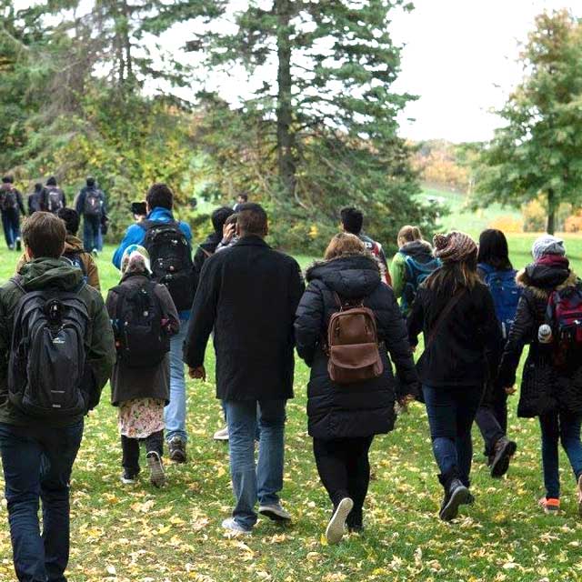 A group of people with backpacks and warm clothing walk through a grassy, tree-lined park in the fall. Some autumn leaves are on the ground, and the environment appears cool and overcast. More people from an international student network can be seen in the background among the trees. A group of people with backpacks, part of an International Student Network, walk through a grassy, tree-lined area on an overcast day.
