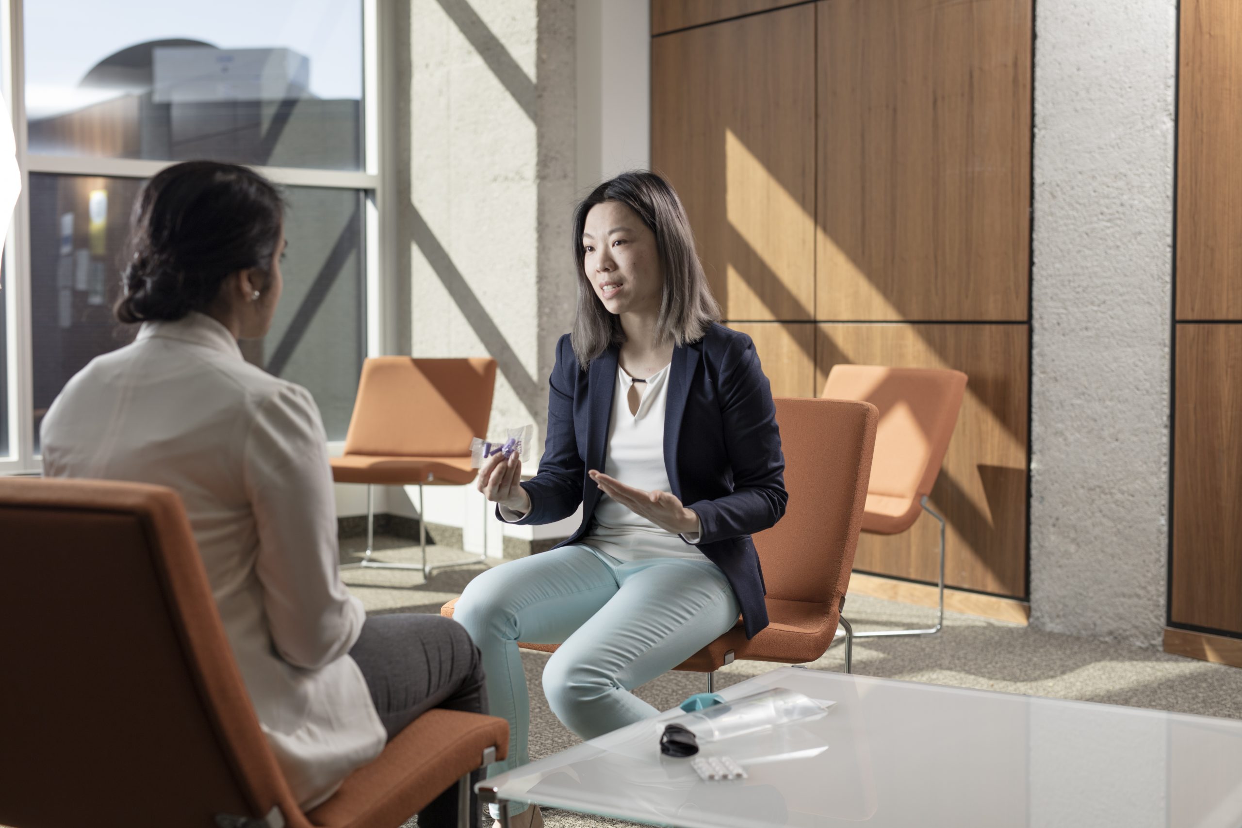 Two women seated in an office lobby, having a conversation. One woman gestures with her hand while holding an object.