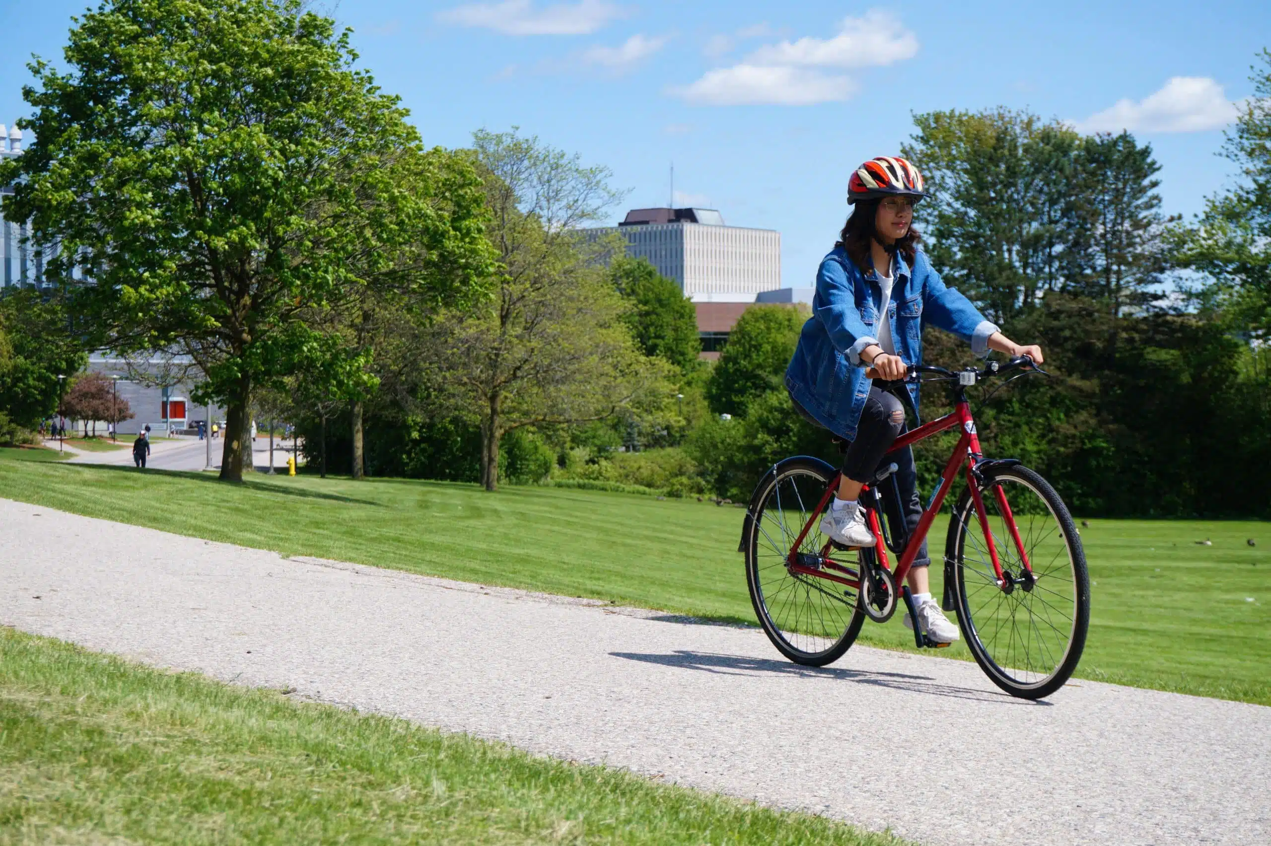 A student rides a red bicycle on a paved path through the park, flanked by green grass and trees, with a building in the background, symbolizing the accessibility of student-run services nearby.