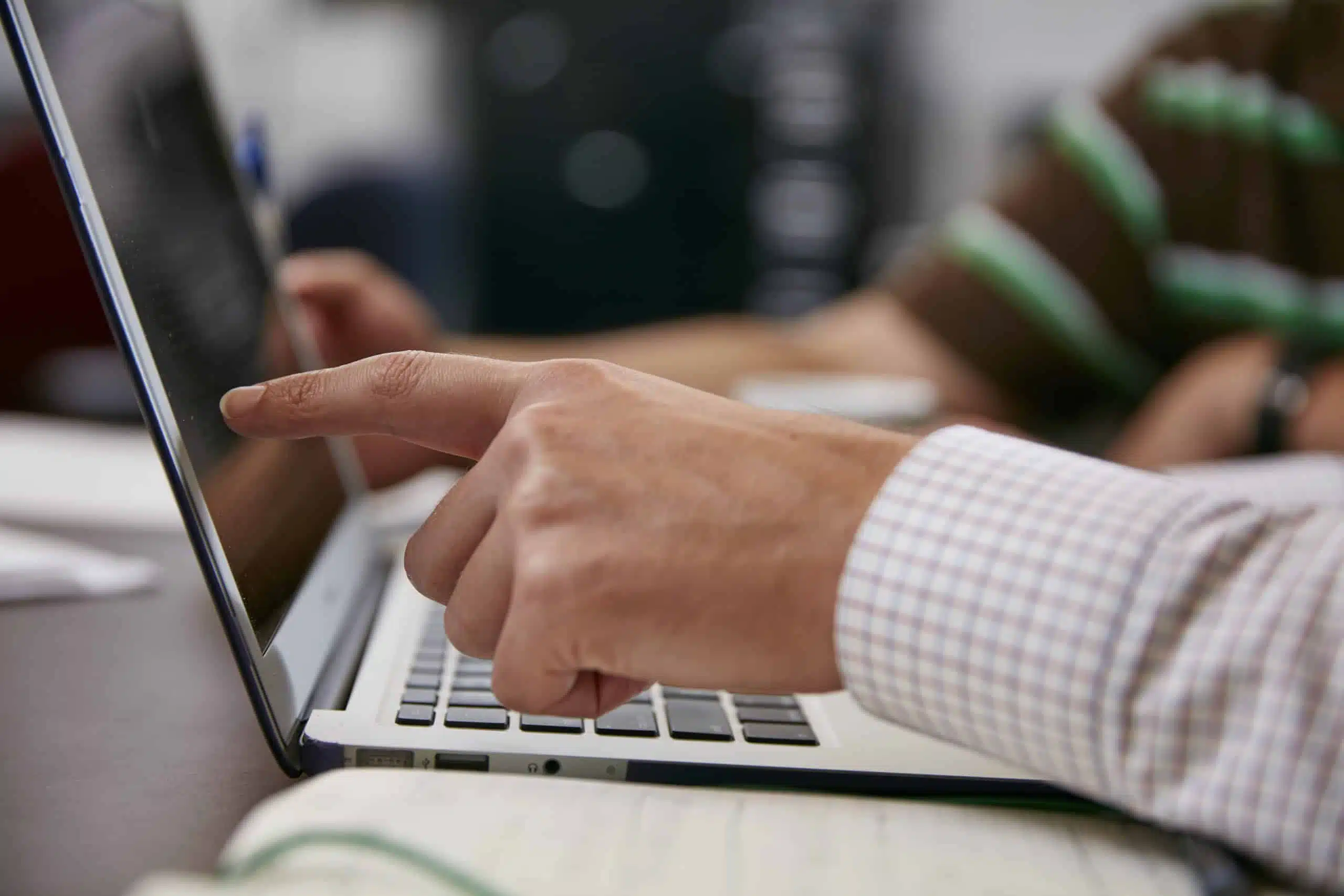 Close-up of a person pointing at a laptop screen, with another person in the background working.