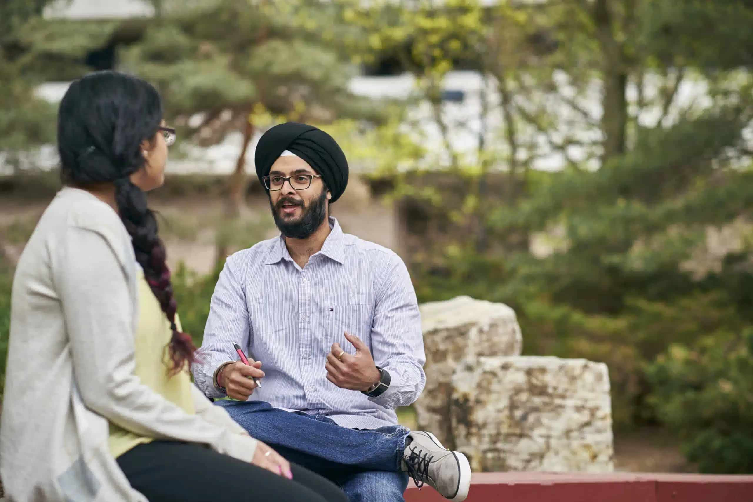 Two individuals sitting outdoors, engaged in conversation. The man is gesturing with his hand while the woman listens. Trees and rocks are visible in the background.