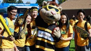A group of students in yellow UWaterloo shirts pose with a King Warrior lion mascot holding a football. The mascot is wearing a striped shirt with "WATERLOO" on it. They are at an outdoor orientation event with a "Let's Make Someday Happen" sign.