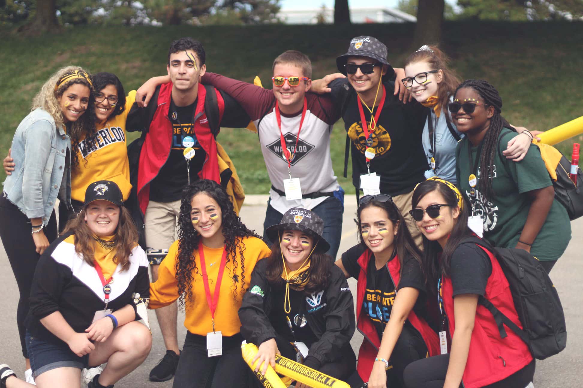 A diverse group of young adults wearing casual clothes and lanyards stand together outdoors, smiling at the camera. Some have face paint and accessories, suggesting they are participating in a team event.