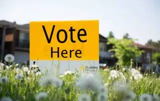 A yellow sign with "Vote Here" written in black is placed in a grassy area with dandelions, indicating a polling station.