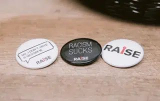 Three buttons are displayed on a wooden surface. The first reads "No, I don't have a nickname," the second says "Racism Sucks," and the third has "Raise" with a tie graphic as the "I." These buttons promote racial advocacy and inclusion, emphasizing the importance of equity for all.