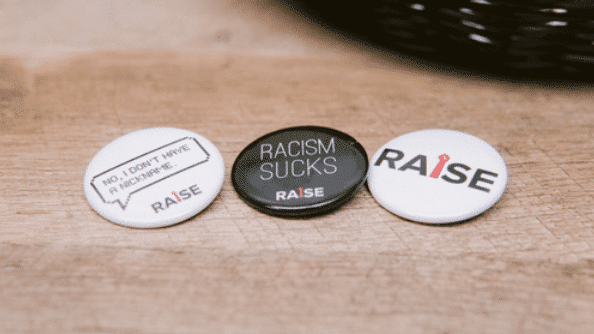 Three round badges are laid out on a wooden surface. The first badge reads “NO, I DON’T HAVE A NICKNAME.” The second badge is black with the message “RACISM SUCKS,” promoting racial advocacy. The third badge displays the word “RAISE” with a tie icon, highlighting the importance of equity in the workplace. Three buttons are displayed on a wooden surface. The first reads "No, I don't have a nickname," the second says "Racism Sucks," and the third has "Raise" with a tie graphic as the "I." These buttons promote racial advocacy and inclusion, emphasizing the importance of equity for all.