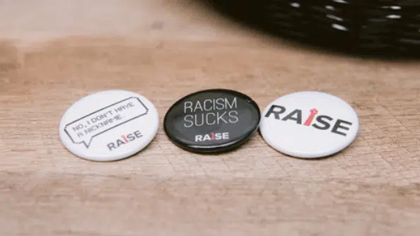 Three buttons are displayed on a wooden surface. The first reads "No, I don't have a nickname," the second says "Racism Sucks," and the third has "Raise" with a tie graphic as the "I." These buttons promote racial advocacy and inclusion, emphasizing the importance of equity for all.