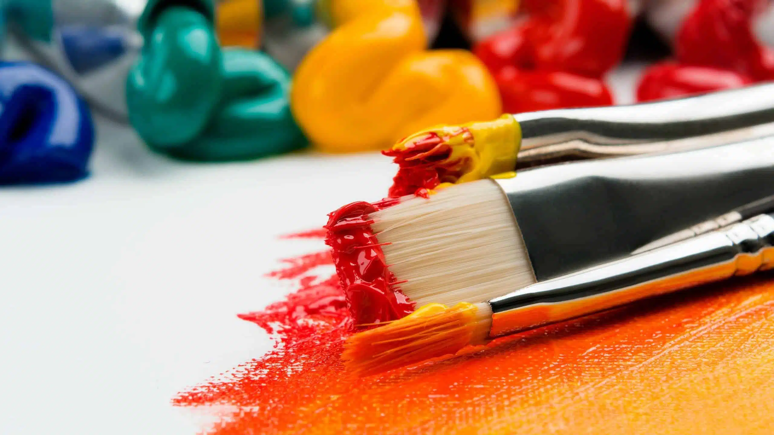 Three paintbrushes with yellow, red, and orange paint on their bristles, with red and orange paint smeared on a white surface. Blobs of various paint colors are in the background, capturing the vibrant energy of a poster run.