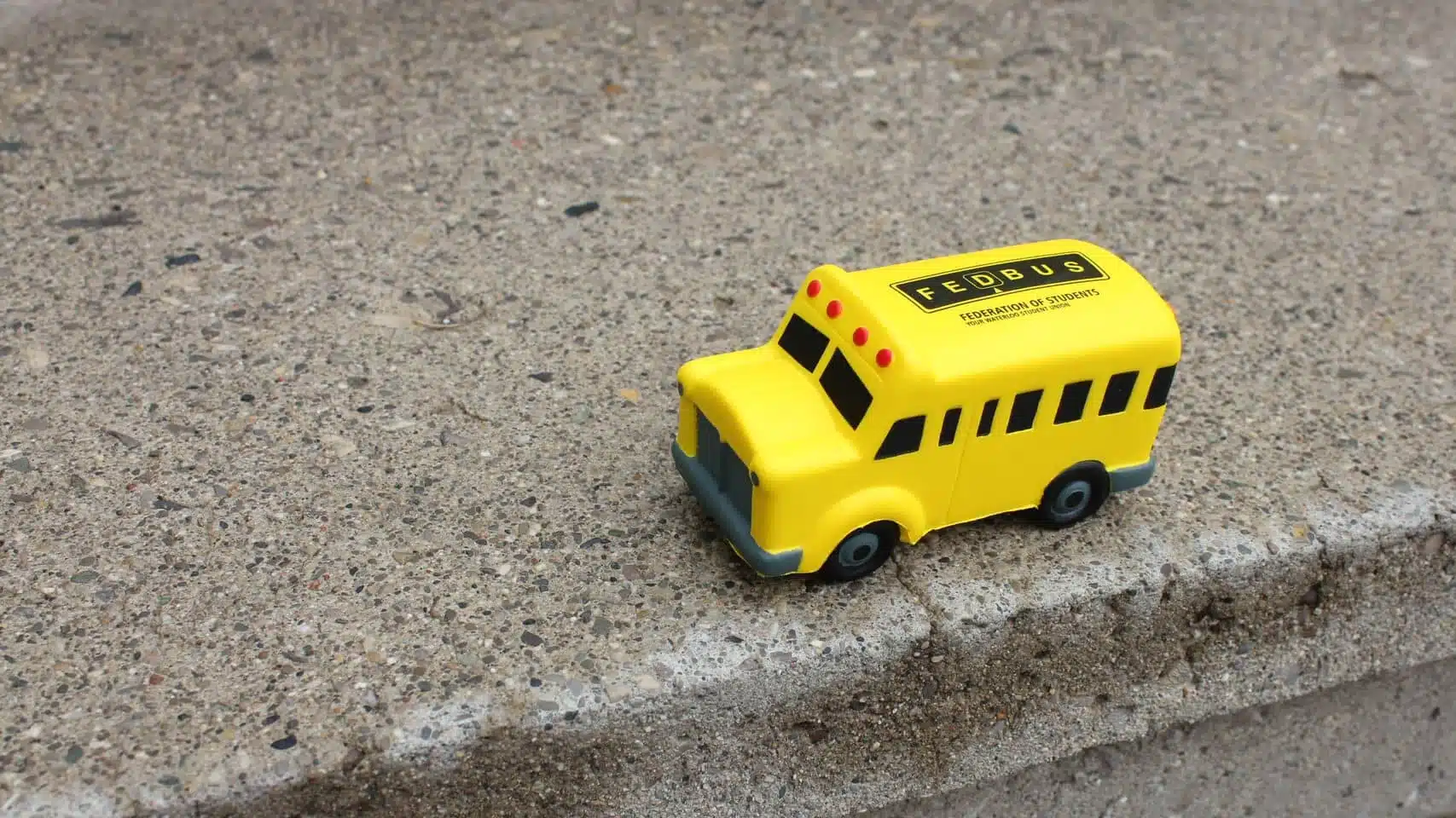 A small yellow toy bus with "FedBus" proudly displayed on its side sits charmingly atop a concrete step.