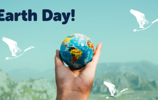 A hand holding a small earth globe against a mountainous backdrop with "Earth Day!" text and white birds in the sky flying in different directions.