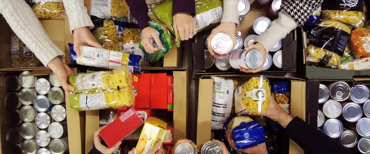 Aerial view of several hands sorting packaged food items, including pasta and canned goods, into cardboard boxes in a retail setting.