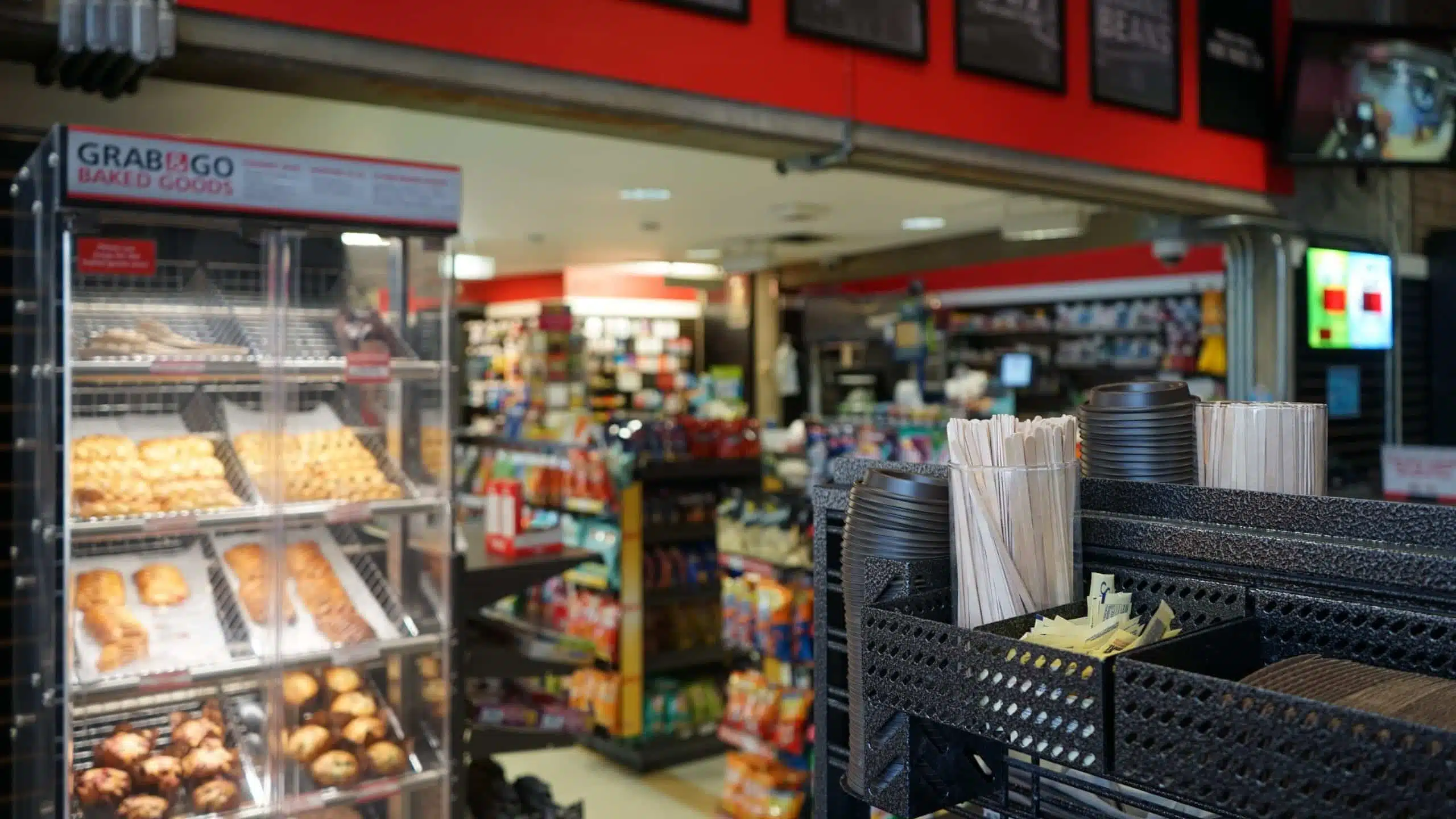 A retail convenience store interior showcasing baked goods on display, various snacks on shelves, and a station with cups, straws, and stirrers in the foreground.