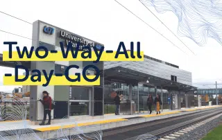 Shot of a train station platform with a modern building in the background. The text "Two-Way-All-Day-GO" is prominently displayed. Passengers, including students who benefit from the reliable GO Train Service, are waiting on the platform, and rail tracks are visible.