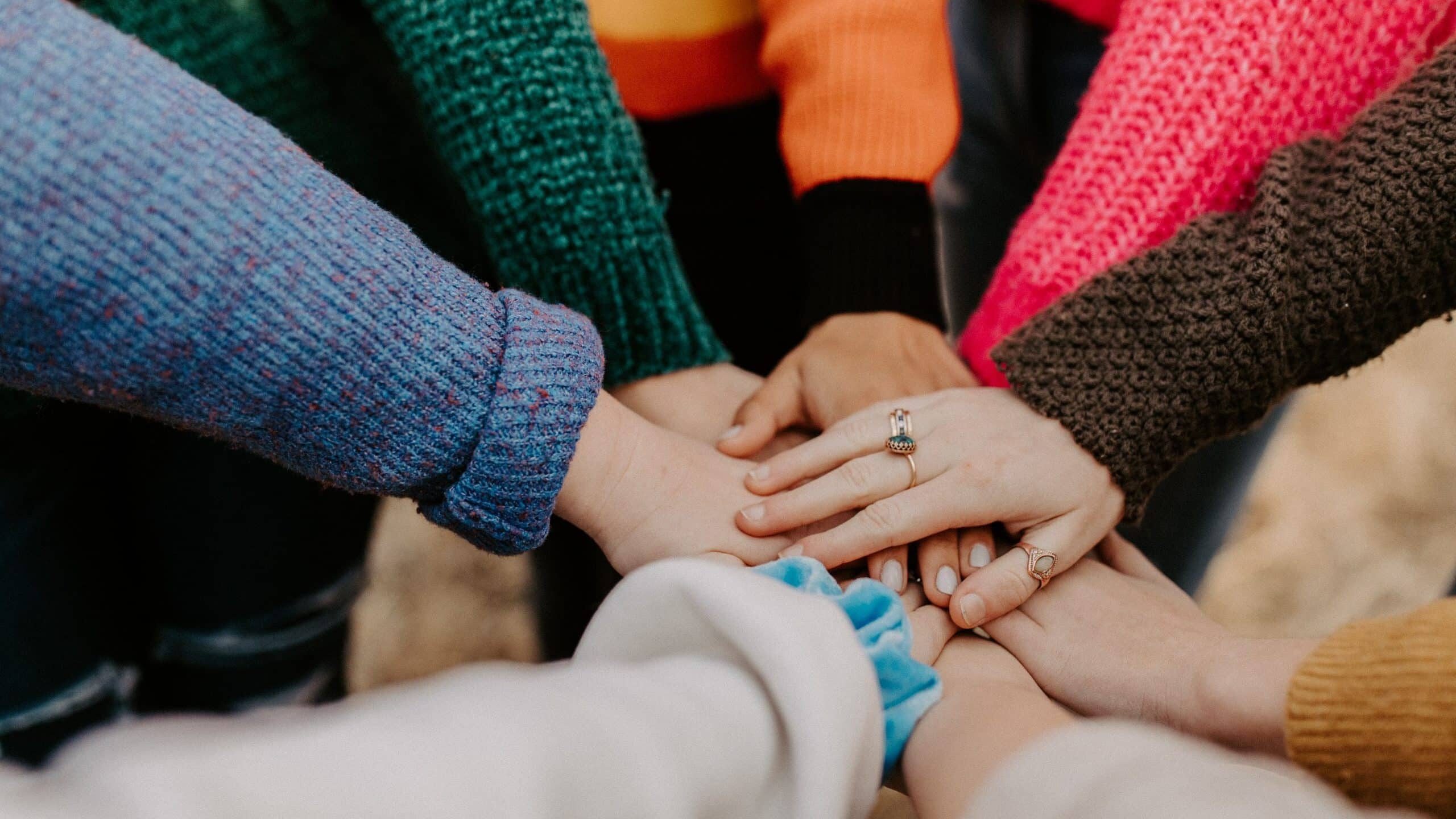 A group of people, wearing colorful sweaters, place their hands together in a huddle.