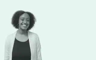 A person with short curly hair is smiling while standing against a plain, light-colored background. They are wearing a dark shirt and a light cardigan, representing the Arts Student Union with pride.