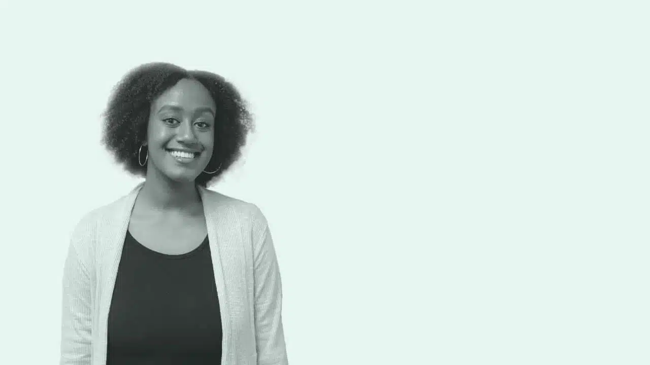 A person with short curly hair is smiling while standing against a plain, light-colored background. They are wearing a dark shirt and a light cardigan, representing the Arts Student Union with pride.