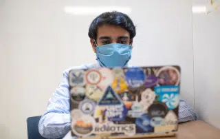 A person wearing a blue mask, seated indoors, is working on a laptop covered in various stickers. Amidst the hustle, a sticker prominently reads "book your vaccination" as a reminder of the ongoing efforts for COVID-19 vaccination in Ontario.