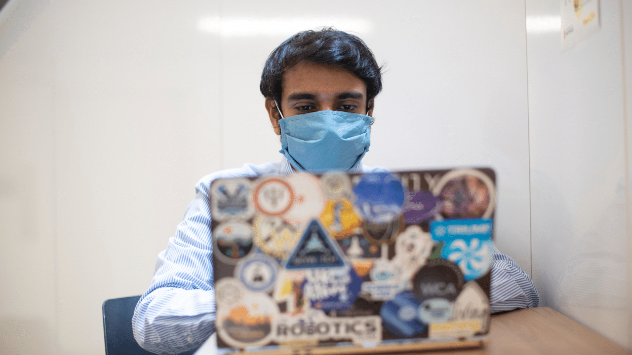A person with dark hair wearing a blue face mask is sitting at a table and working on a laptop covered in various colorful stickers. The background is a plain, light-colored wall, emphasizing the importance of health measures while you book your COVID-19 vaccination in Ontario. A person wearing a blue mask, seated indoors, is working on a laptop covered in various stickers. Amidst the hustle, a sticker prominently reads "book your vaccination" as a reminder of the ongoing efforts for COVID-19 vaccination in Ontario.