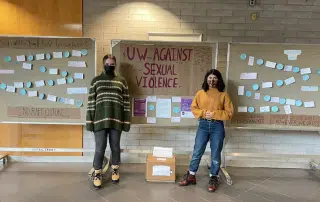 Two individuals stand in front of display boards with messages against sexual violence. The board in the center reads "UW - Against Sexual Violence" surrounded by informational posters and notes, emphasizing the ongoing fight to eliminate violence against women.