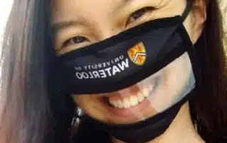 A person with long dark hair is wearing a black face mask with a clear plastic window over the mouth area for mask accessibility, and the mask has "University of Waterloo" written on it.