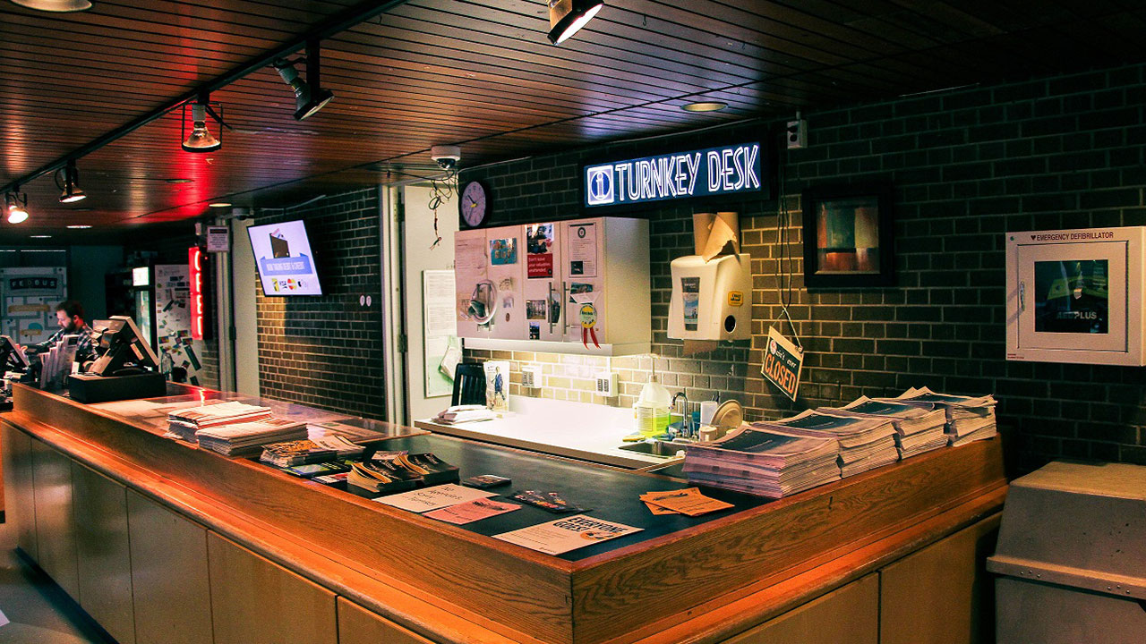 A reception desk labeled "O'Turkey Desk" with pamphlets, papers, and a phone. The area is lit with overhead lights and there are various signs and pictures on the wall behind the desk.