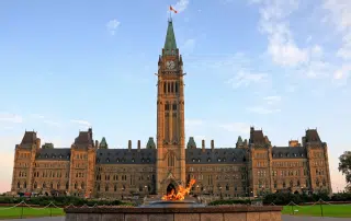 The image shows the Canadian Parliament Buildings in Ottawa, with the Peace Tower and a prominent central clock. The Centennial Flame is burning in the foreground, symbolizing unity and history across every province.