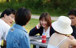 A group of five students sit around an outdoor picnic table. One person has a can of Coca-Cola in front of them. They are talking and engaging with each other, likely supporting international students among them. Outdoor greenery is visible in the background.