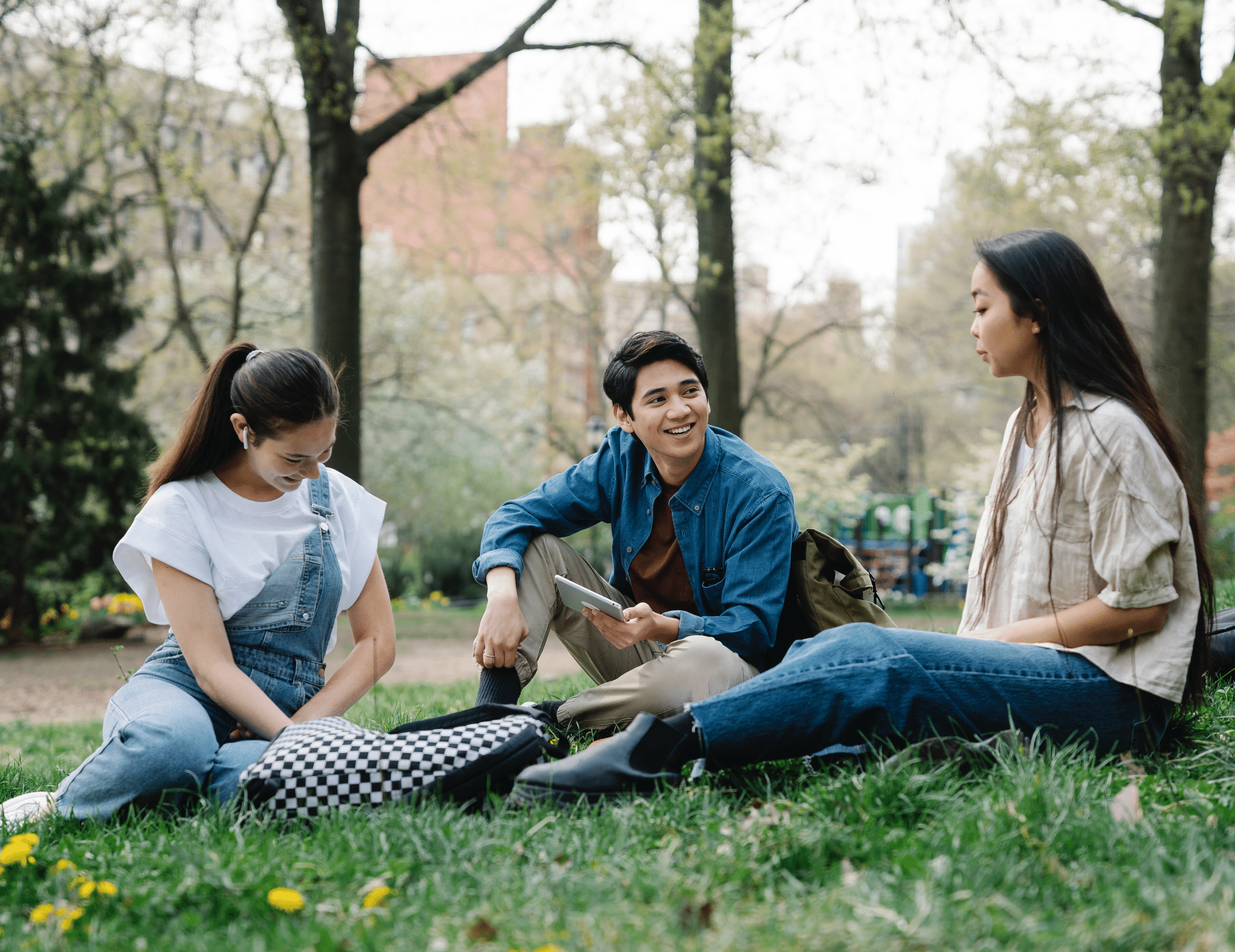 Three young people are sitting on the grass in a park, engaged in conversation. One person is holding a tablet, and they are all smiling. Trees and buildings can be seen in the background, suggesting an urban park setting—an ideal scene for WUSA Washington's latest community news snapshot.