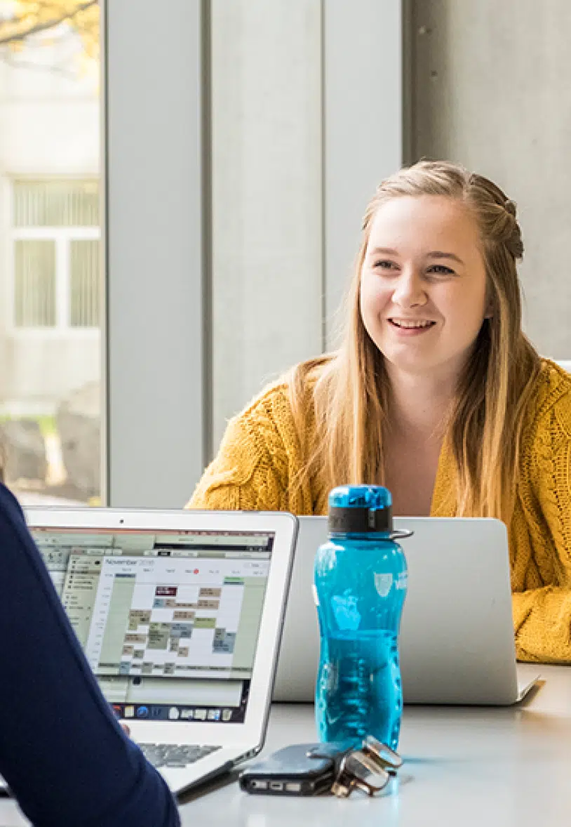 A young woman with long blonde hair, dressed in a yellow sweater, smiles while working on a laptop. She sits across from another person using a laptop. A blue water bottle, keychain, and glasses are on the table between them. The background has large windows.