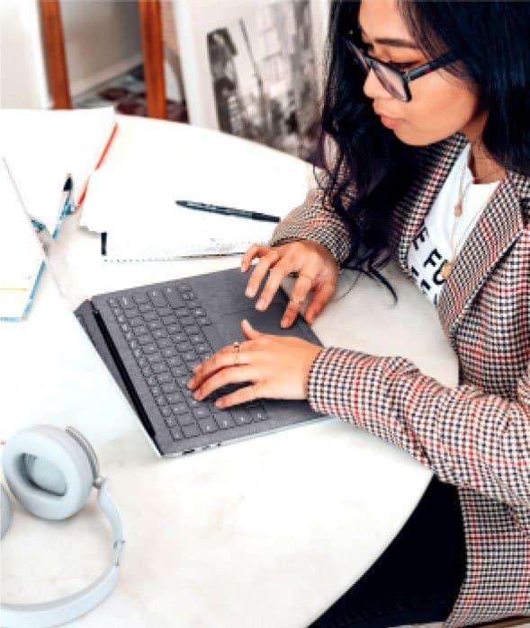 A person with long dark hair and glasses types on a laptop at a white table. The table has notebooks, a pen, a smartphone, and headphones. The person is wearing a plaid blazer and white top, focused on their work in a bright, modern setting.