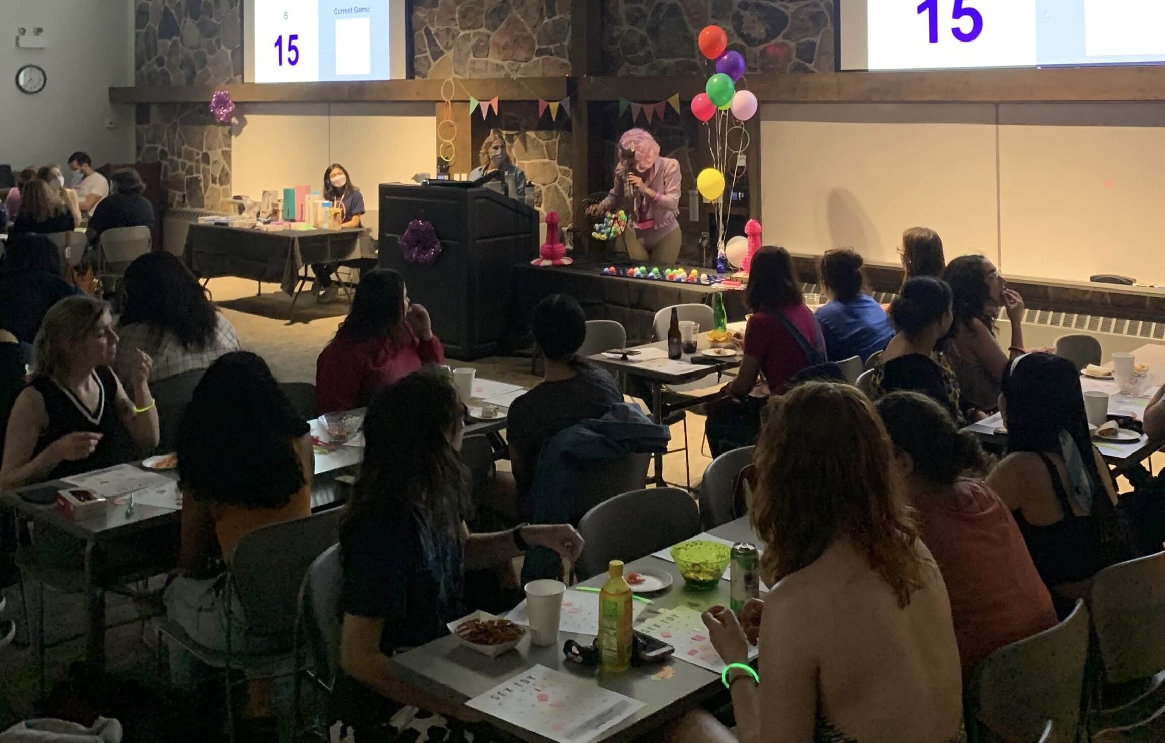 A dimly lit room with people seated at tables, watching a person dressed in a colorful outfit and wig performing on a stage. The stage, decorated with balloons and a small table, sets the scene for one of Catherine's Top WUSA Events. Two screens display the number 15. The ambiance is casual and festive.