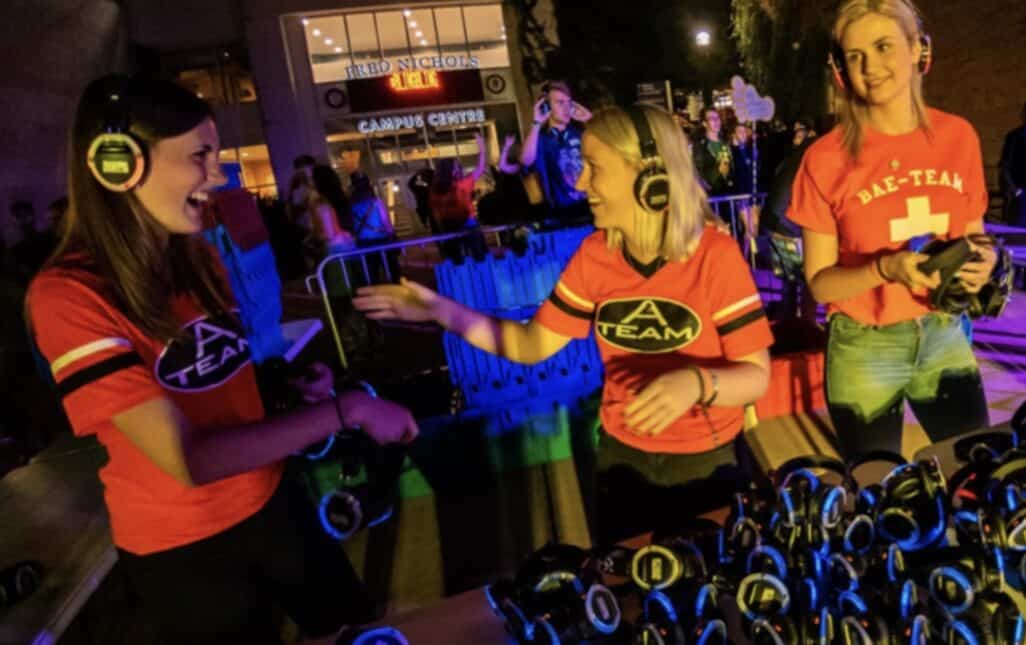 At a top event hosted by WUSA events, three women wearing red shirts are running a headphone booth at a nighttime gathering. They are smiling and equipped with headphones, interacting with attendees. In the background, people are mingling near a building labeled "Campus Center." The area is illuminated with colorful lights.