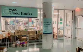 Photo of the WUSA Used Books entrance with a banner reading "WUSA Used Books." Boxes filled with secondhand books are placed on tables near the entrance. A sign on a pillar advertises "Save 20% on Textbooks." Behind the entrance, shelves of books are visible.